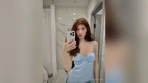 Redheaded woman in a light blue satin slip dress taking a mirror selfie in a modern marble bathroom.