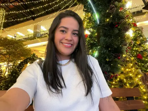 Smiling woman in a white t-shirt sitting by a glowing Christmas tree in a festive holiday setting.