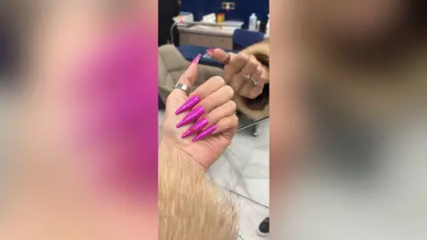 Close-up of long pink stiletto nails and silver rings on hands held in a bright salon setting.