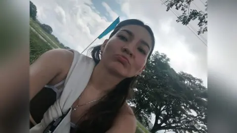 Young woman in a white sleeveless top smiling outdoors against a backdrop of green trees and a cloudy sky.