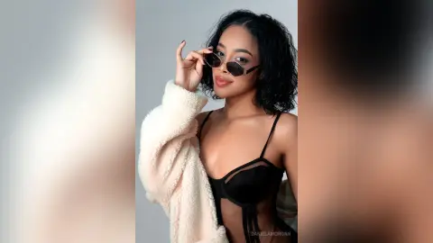 DanielaMorgan poses in a black lace bra and sunglasses, wearing a white fuzzy jacket over her shoulders.