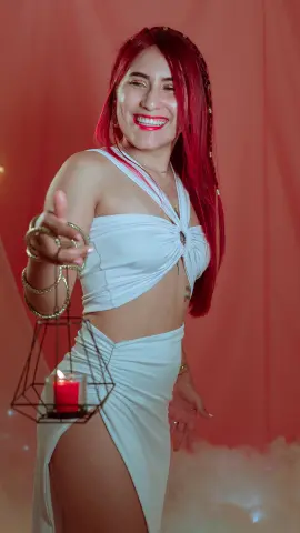 Smiling woman with red hair in a white cutout outfit holding a candle against a pink background.