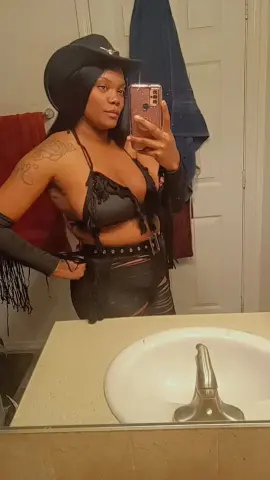 Confident woman in black western cowgirl outfit and hat taking a mirror selfie in a bathroom.