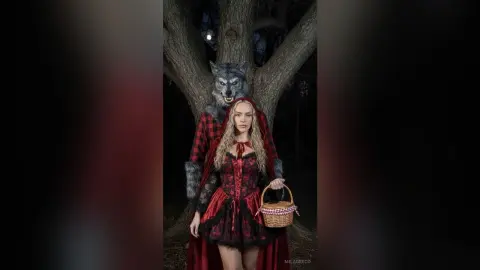 MilaGreco poses in a red and black lace outfit with a matching hooded cape, holding a small basket while standing in front of a large tree and a wolf head.