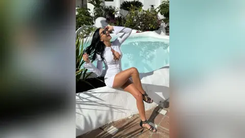 Smiling woman in a white long-sleeve mini dress relaxing by a bright turquoise swimming pool on a sunny day.