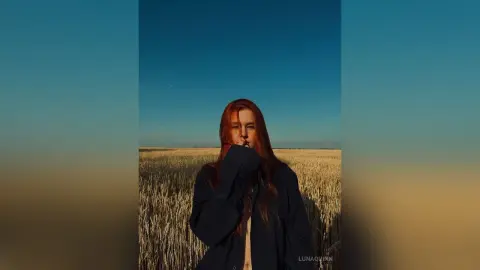 Redheaded woman in a dark navy shirt standing in a golden wheat field under a clear blue sky at sunset.