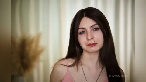 Young woman with long brown hair and nose piercing wearing a pink tank top in a bright indoor setting.