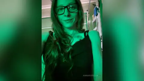 Smiling woman with long hair and glasses wearing a black camisole in moody green lighting.
