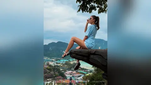 AnaMathers sits on a large rock overlooking a tropical landscape, wearing a light blue t-shirt and sunglasses.