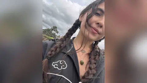 Smiling woman with long dark braids wearing a black zip-up jacket outdoors under a cloudy sky.
