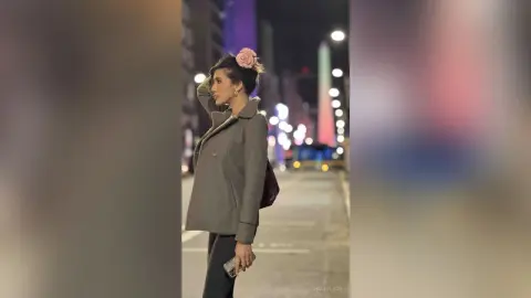 HaleyLyon poses on a city street at night, wearing a grey button-up jacket and dark pants with a pink flower in her hair.