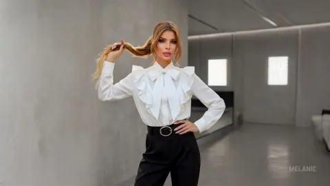Elegant woman in a white ruffled bow blouse and black trousers posing in a modern minimalist interior.