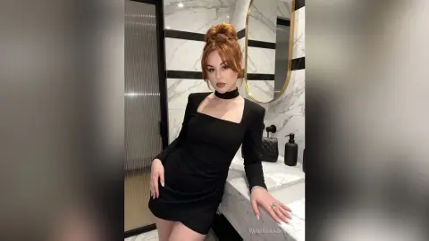 Elegant redhead woman in a chic black long-sleeve dress posing in a modern marble bathroom.