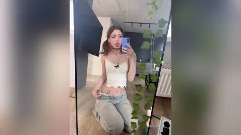 Young woman in a white lace camisole and light denim jeans taking a selfie in a modern minimalist room.
