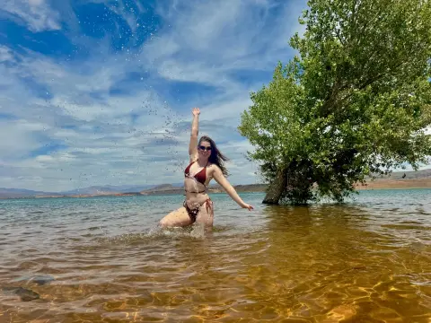 Happy woman in a bikini splashing in a clear blue lake under a sunny sky with green trees.