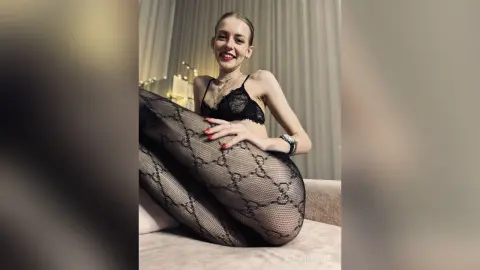 Smiling woman in black lace lingerie and patterned fishnet tights relaxing on a sofa in a dimly lit room.