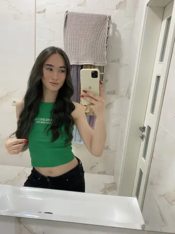 Young woman in a green tank top taking a mirror selfie in a modern marble bathroom.