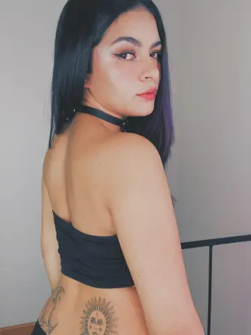 Young woman with long dark hair wearing a black tube top and choker in a minimalist indoor setting.