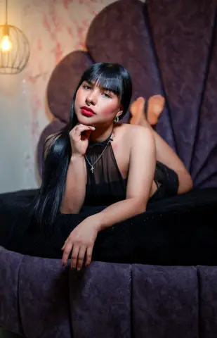 Glamorous woman in a black sheer top relaxing on a purple velvet shell chair in a moody, elegant setting.