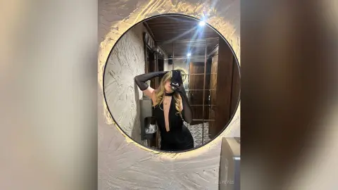 Glamorous woman in a chic black evening dress taking a mirror selfie in a modern, dimly lit bathroom.