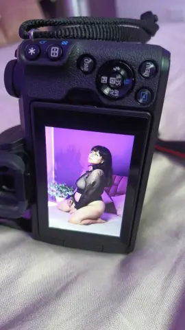 LuluPup poses naked in a black lace bodysuit while kneeling on a white surface against a purple background.