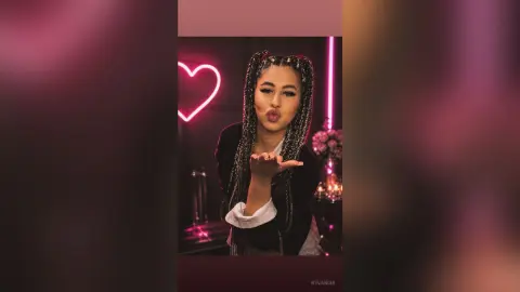 Smiling woman with long braids in a black blazer blowing a kiss near a pink neon heart sign.
