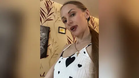 Smiling woman in a white polka dot tank top posing in a bright room with floral wallpaper.