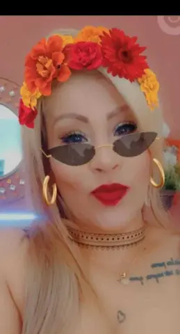 Blonde woman with red lipstick and floral crown wearing gold hoop earrings and sunglasses in a bright setting.