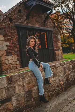 Young woman in a black long-sleeve top and light blue denim jeans sitting on a stone wall by a rustic brick house.