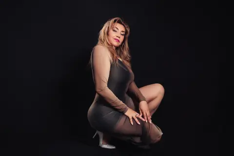 Confident woman in a sheer grey bodysuit posing against a black background in a moody studio setting.