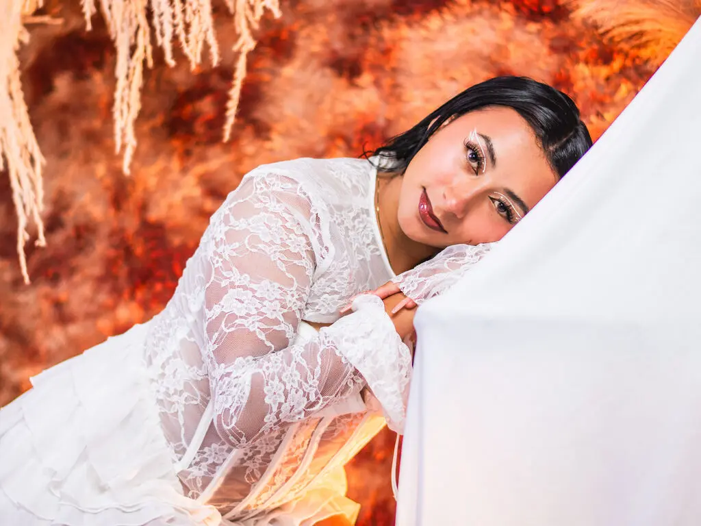Elegant woman in a white lace dress leaning against a white surface against a warm, textured orange background.
