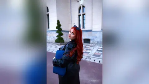 Red-haired woman in a black jacket holding a blue folder walking through a historic city plaza.