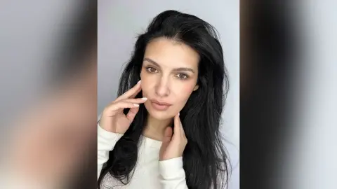 Portrait of a woman with long dark hair wearing a white ribbed sweater against a neutral studio background.