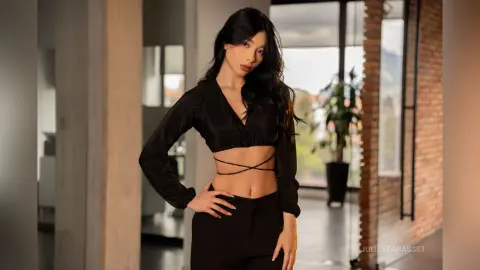 Stylish woman in a black long-sleeve crop top and trousers posing in a modern, sunlit loft apartment.