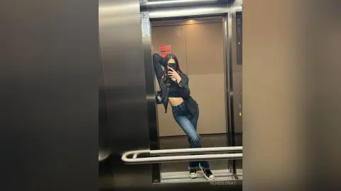 Stylish woman in a black crop top and jeans taking a mirror selfie in a modern elevator.