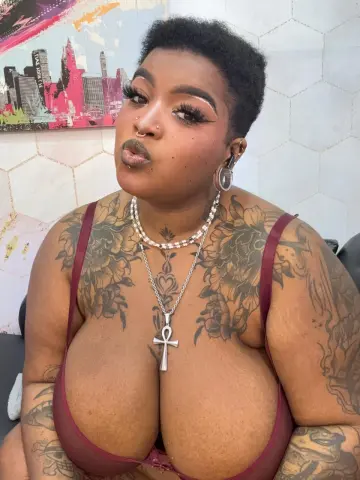 Tattooed woman in a burgundy camisole wearing silver jewelry and an Ankh necklace in a bright room.