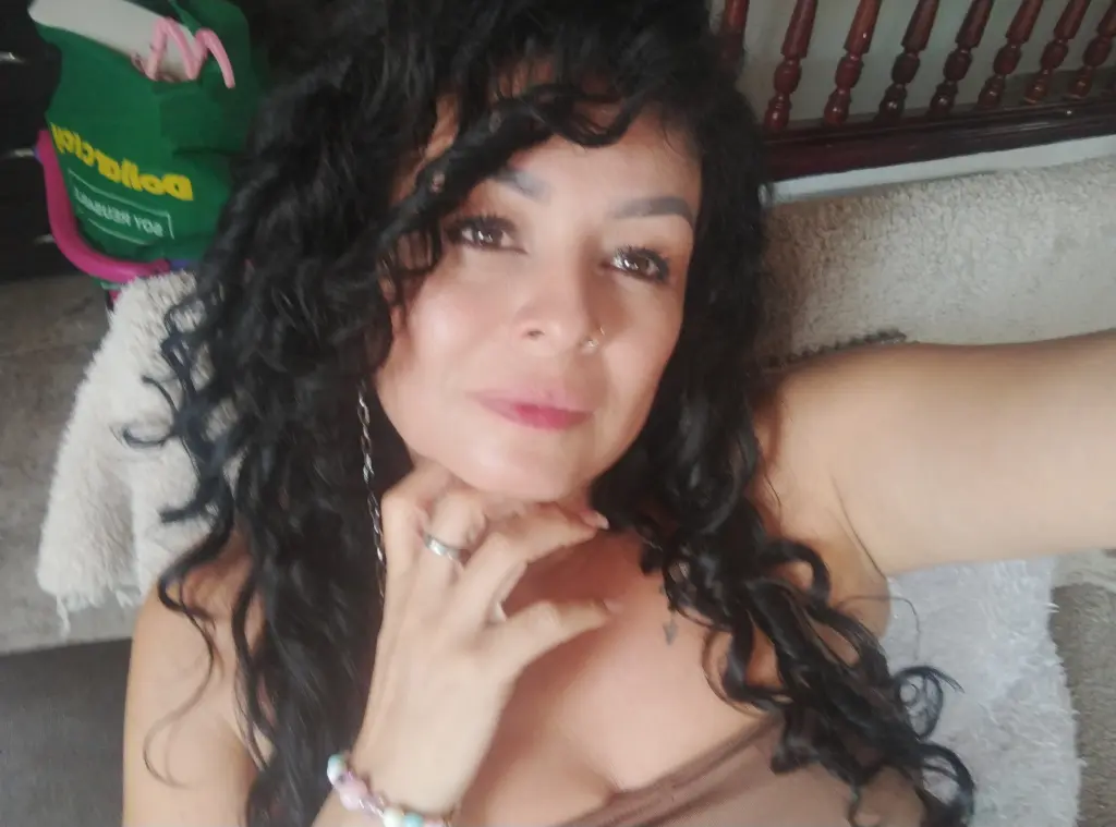 Woman with curly dark hair wearing a black top and pearl necklace in a soft, indoor setting.