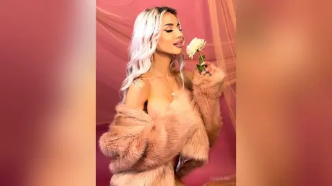 Glamorous blonde woman in a pink faux fur coat holding a white rose against a soft pink backdrop.