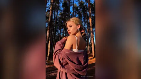 Blonde woman in a mauve cardigan posing in a sunlit pine forest during golden hour.