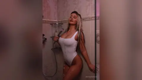 Anita poses in a white bodysuit while standing in a tiled shower with wet skin.