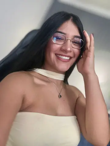Smiling young woman with glasses in a cream halter top posing in a cozy indoor setting.