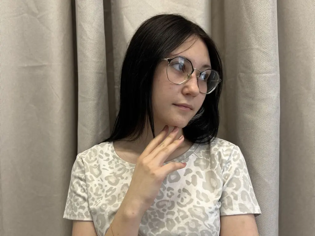 LanelleHiga wearing glasses and a white leopard print t-shirt, posing with her hand against her neck against a neutral curtain background.