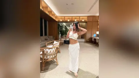 Young woman in a white crop top and long skirt taking a mirror selfie in a modern, bright living room.