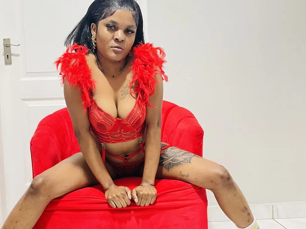 BiancaGreene sits on a red chair wearing a red lace bra and matching red thong, showing her tattoos and dark hair.