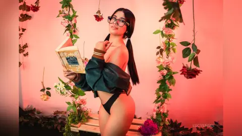 Young woman in a green jacket holding a book amidst pink roses in a romantic, soft-lit studio setting.