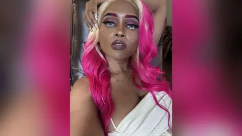 BossyBella — BossyBella poses with her long pink and blonde hair, wearing a white top and gold chain, looking directly at the camera.