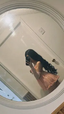 Young woman in a pink ruffled mini dress taking a mirror selfie in a bright room.