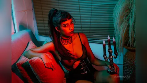 LilithBrooks poses in black lace lingerie and a choker, sitting next to a lit candelabra in a dimly lit room.
