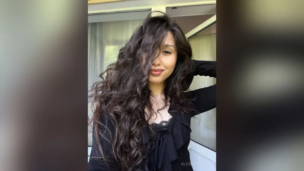 Woman with long wavy brunette hair wearing a black top in a bright, natural indoor setting.