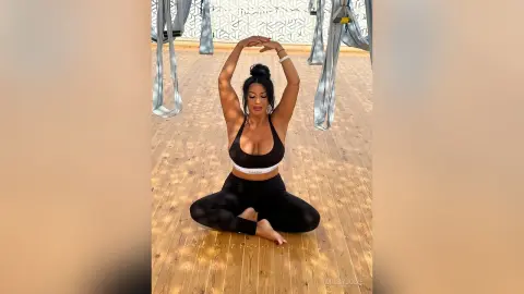 MissyJolie sits cross-legged on a wooden floor wearing a black sports bra and leggings with her hands raised above her head.